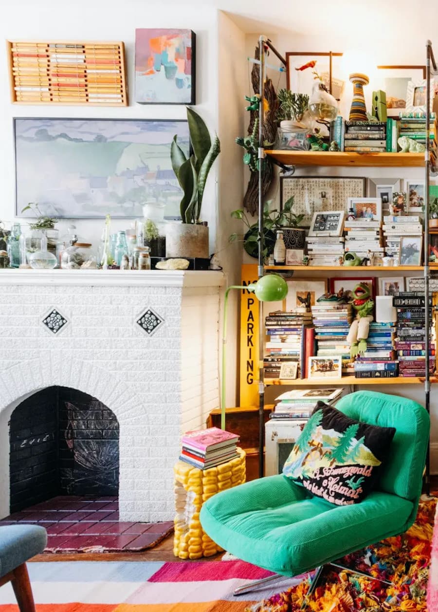 Living con una chimenea de ladrillo blanca con muchos frascos de vidrio sobre ella y cuadros en la pared de la misma. Al costado hay una poltrona verde con un cojín de colores, y detrás de ella, un estante lleno de adornos, libros, plantas y recuerdos.