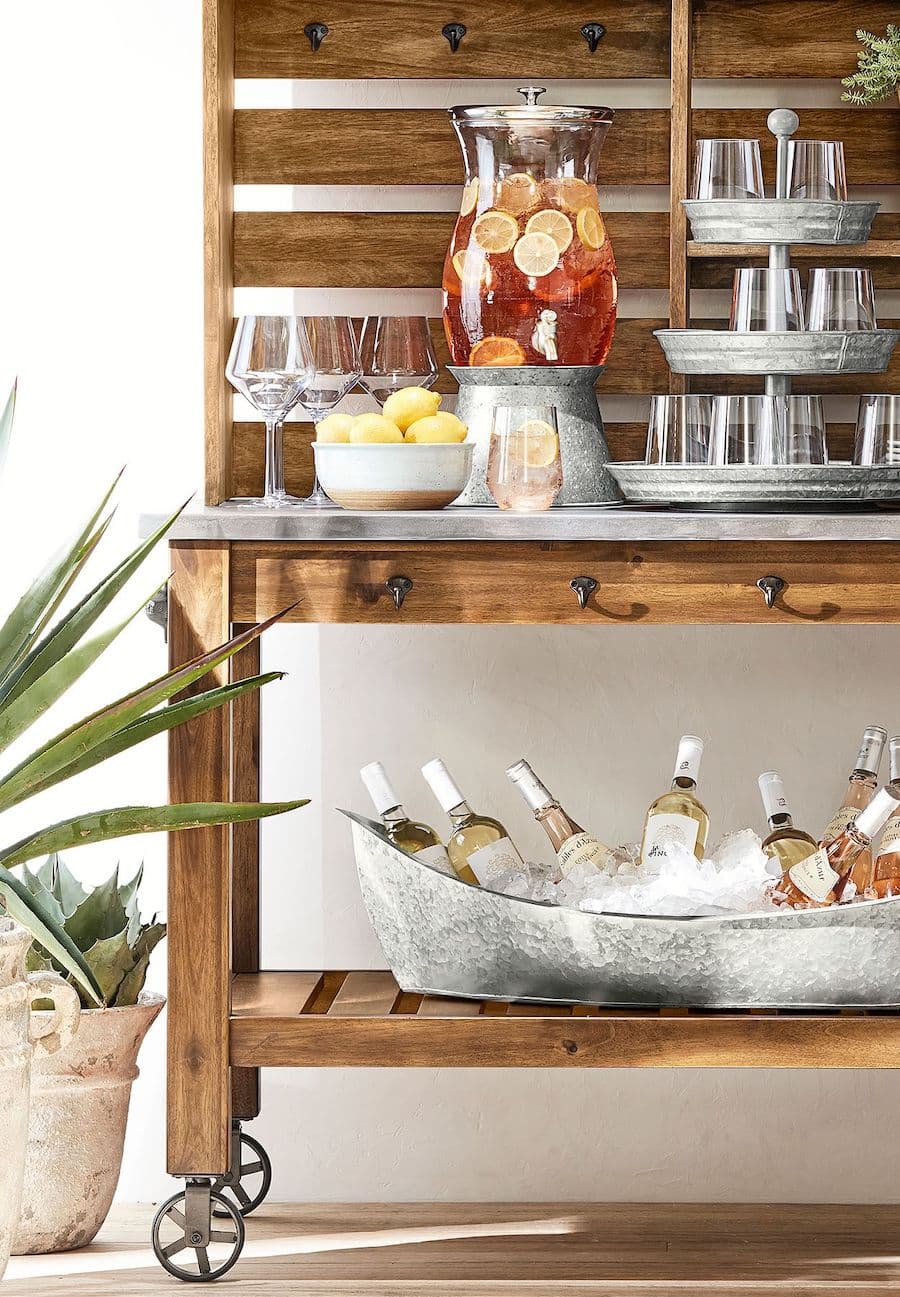 Carro bar de madera de dos niveles. Arriba tiene vajilla y en la parte inferior una cubeta con hielo y botellas. Junto al mueble hay plantas