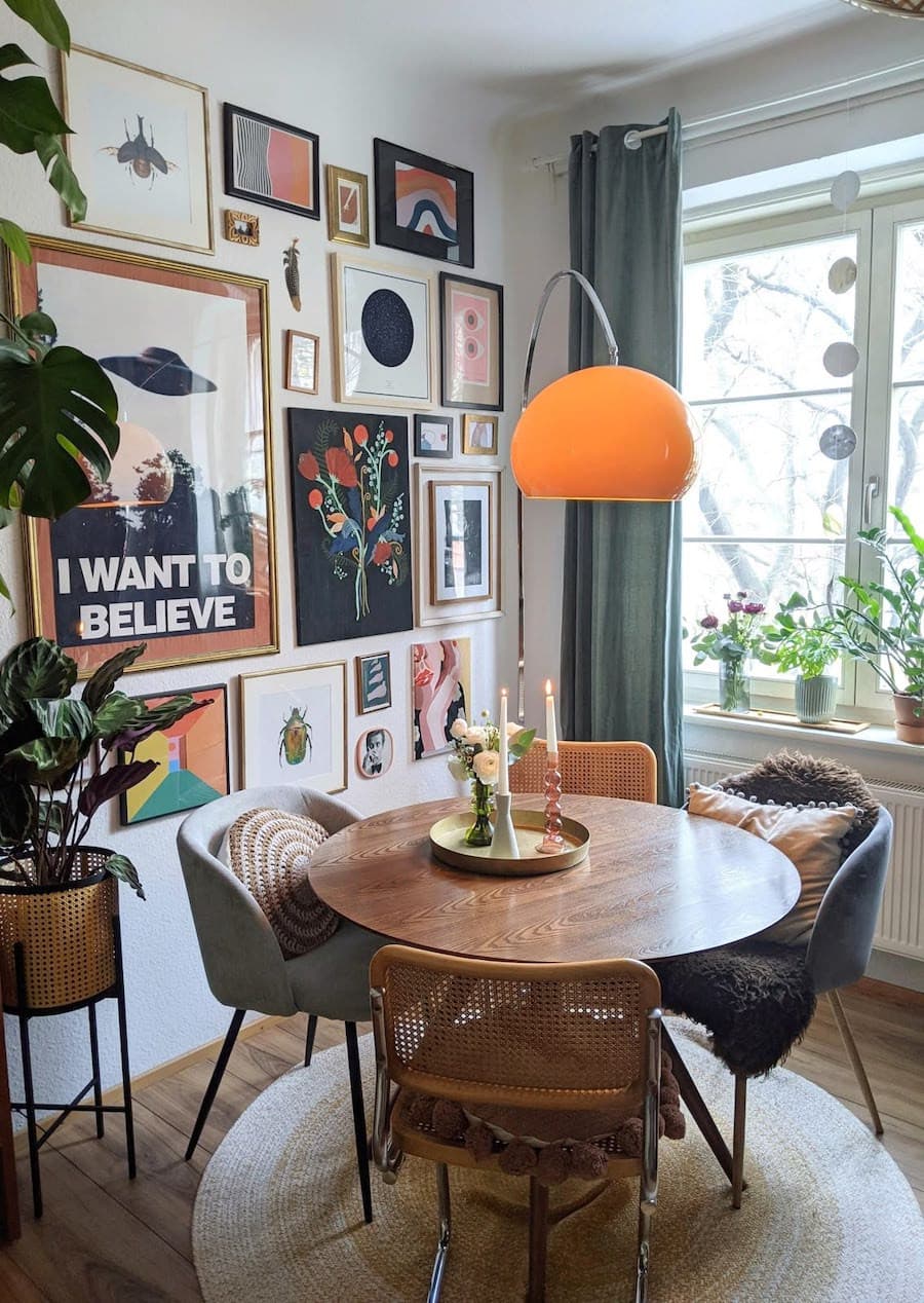 Comedor de estilo cluttercore con una galería de cuadros en la pared, una mesa de comedor redonda de madera, sillas de comedor de distintos estilos y colores, y una lámpara de pie escultural con pantalla anaranjada.