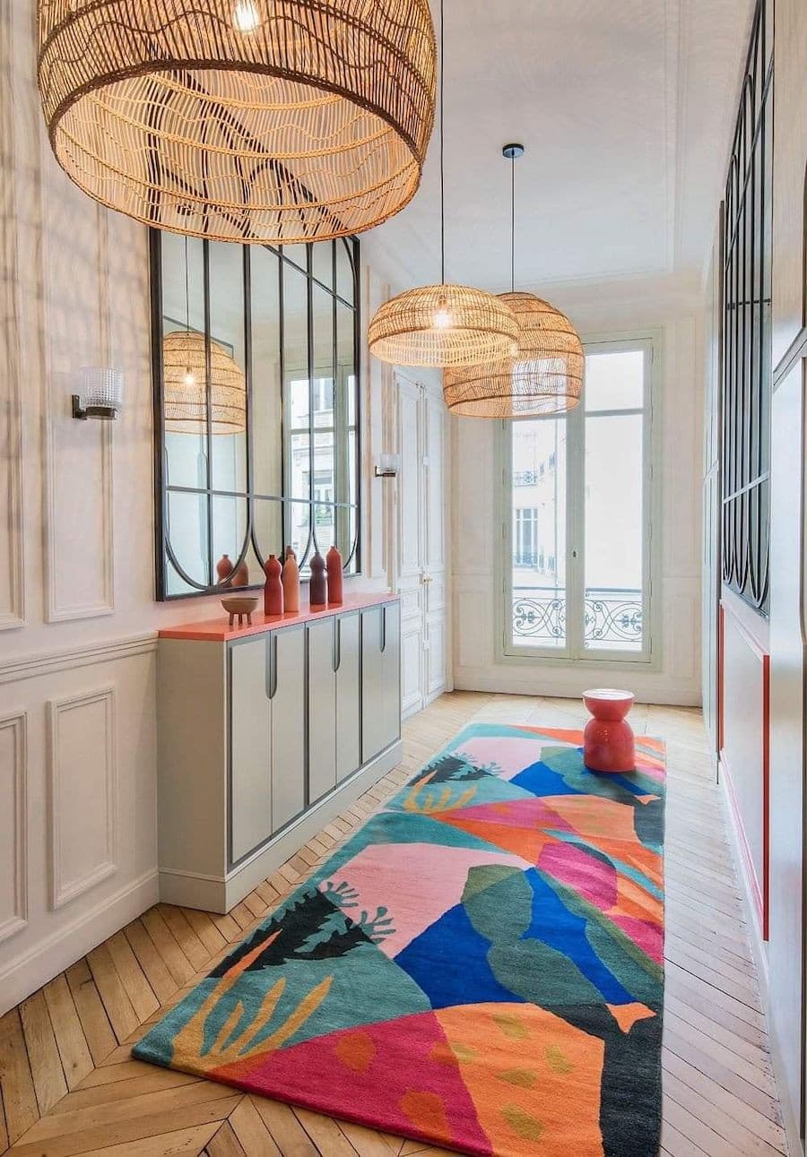 Pasillo con una alfombra de formas geométricas abstractas y colores vibrantes. Desde el techo cuelgan 3 grandes lámparas de libras naturales.
