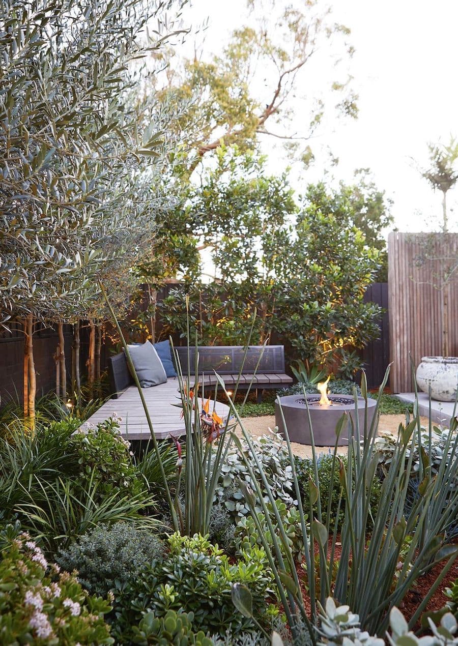 Gran terraza en un jardín, rodeada de plantas y árboles. Muebles a medida, de madera oscura, con cojines grises y un fogón encendido al centro.