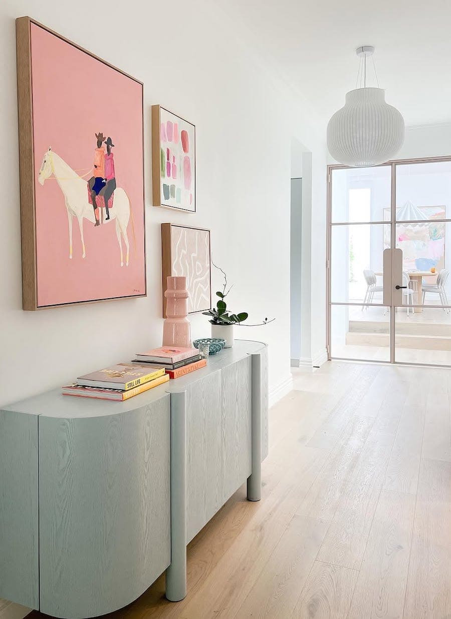 Pasillo con arrimo moderno color celeste. Muros blancos con cuadro rosado. Pisos de madera clara y puerta de vidrio con marco metálico blanco.