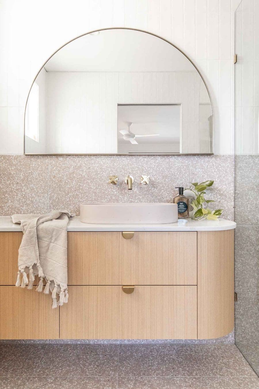 Baño moderno con la mitad del muro con azulejo estilo terrazo en color gris. La parte de arriba tiene azulejos blancos. Gran espejo de arco con marco plateado. Vanitorio de madera con tiradores curvos, en color dorado.