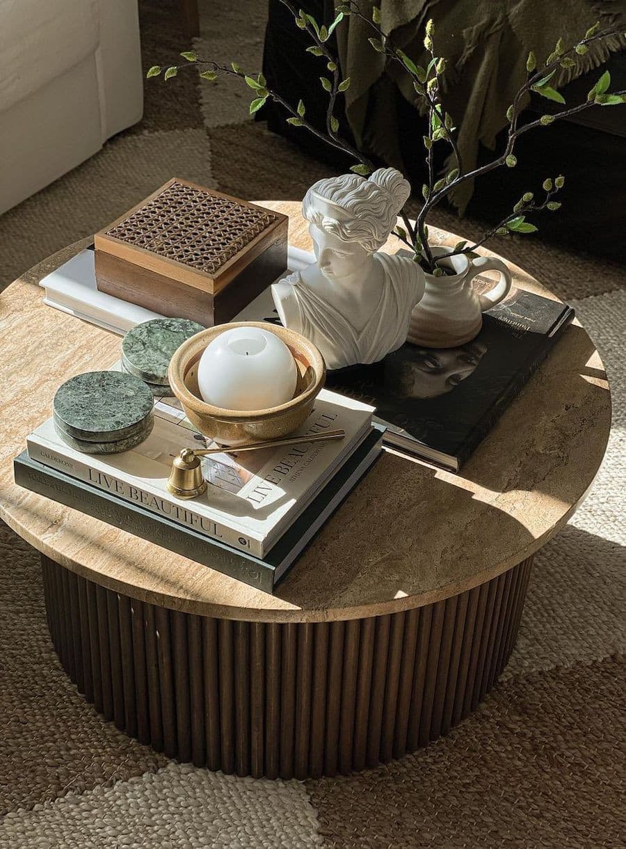 Mesa de centro redonda, de madera. Sobre ella hay libros y revistas, Sobre ellos cajas decorativas, un porta incienso, una escultura de un busto, plantas y una caja de madera