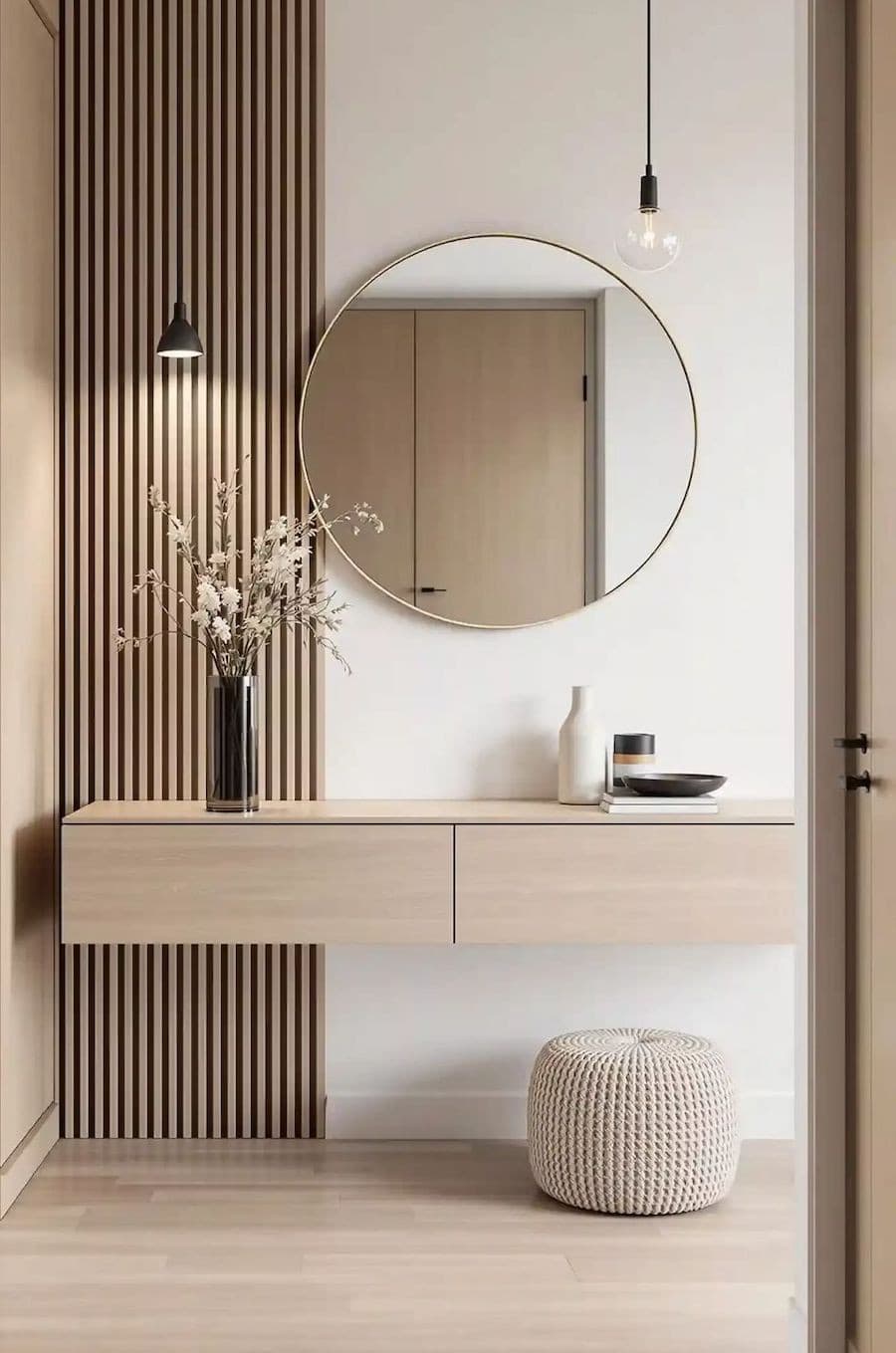 Entrada moderna con un mueble flotante de madera clara que sirve como arrimo. Sobre él hay un florero con flores rosadas y 2 jarrones pequeños al otro costado. También hay un gran espejo redondo, un panel de madera y un apliqué de pared negro.