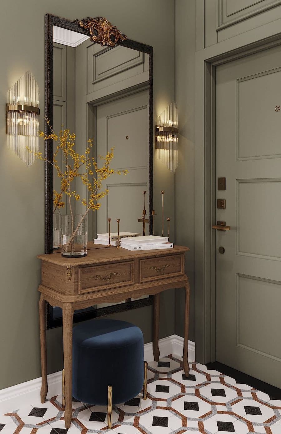 Entrada con un mueble arrimo de madera, un espejo rectangular de cuerpo completo y marco negro, 2 apliqués de pared dorados a los costados, sobre la mesa hay un florero de vidrio con flores amarillas, una pila de libros, y debajo un pouf de terciopelo.