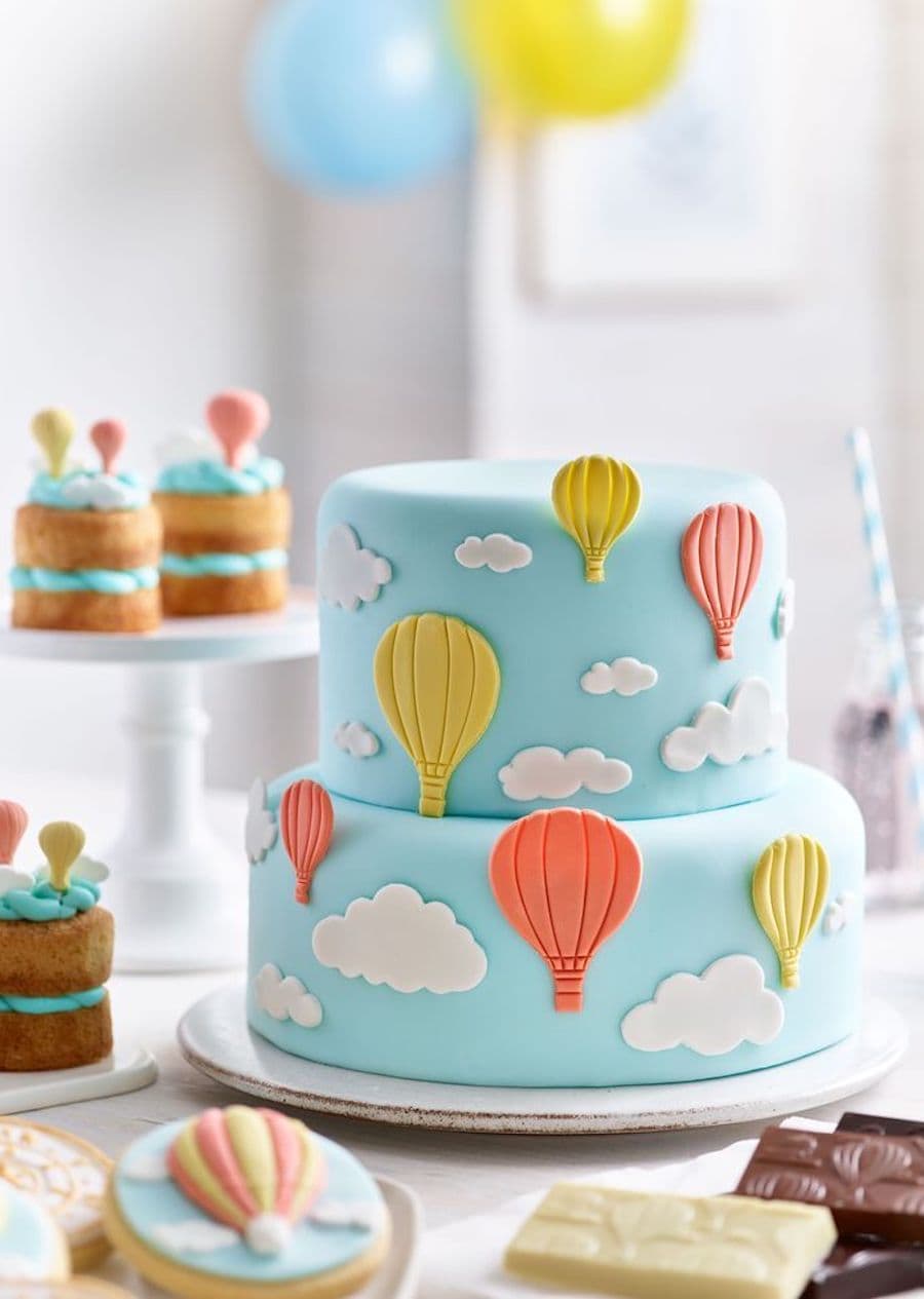 Detalle de una mesa de baby shower con una torta de 2 pisos de nubes y globos aerostáticos amarillos y naranjas, sobre un fondo celeste.