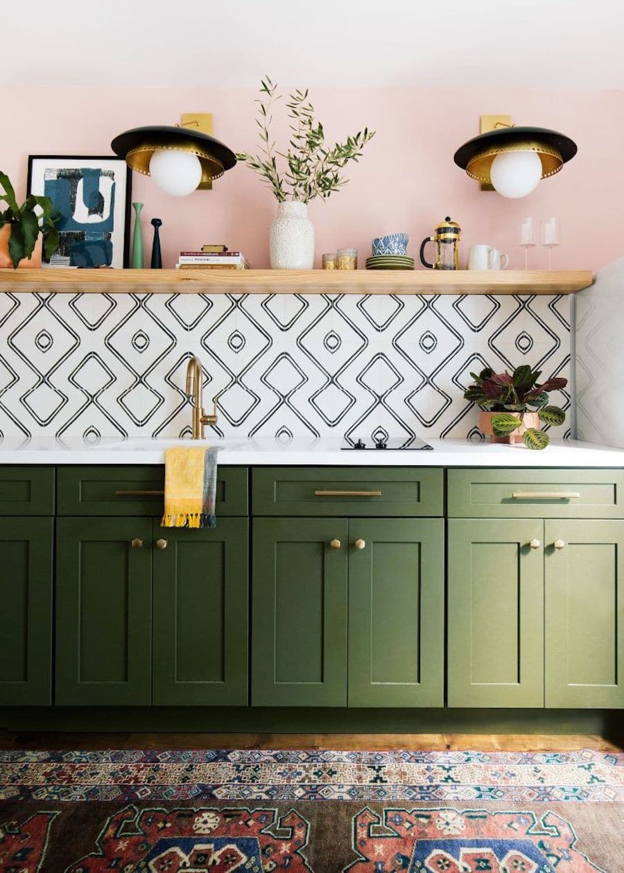 Cocina con muebles inferiores verde, muros rosados, azulejos con un diseño en blanco y negro, y una repisa de madera con decoraciones, menaje y copas.