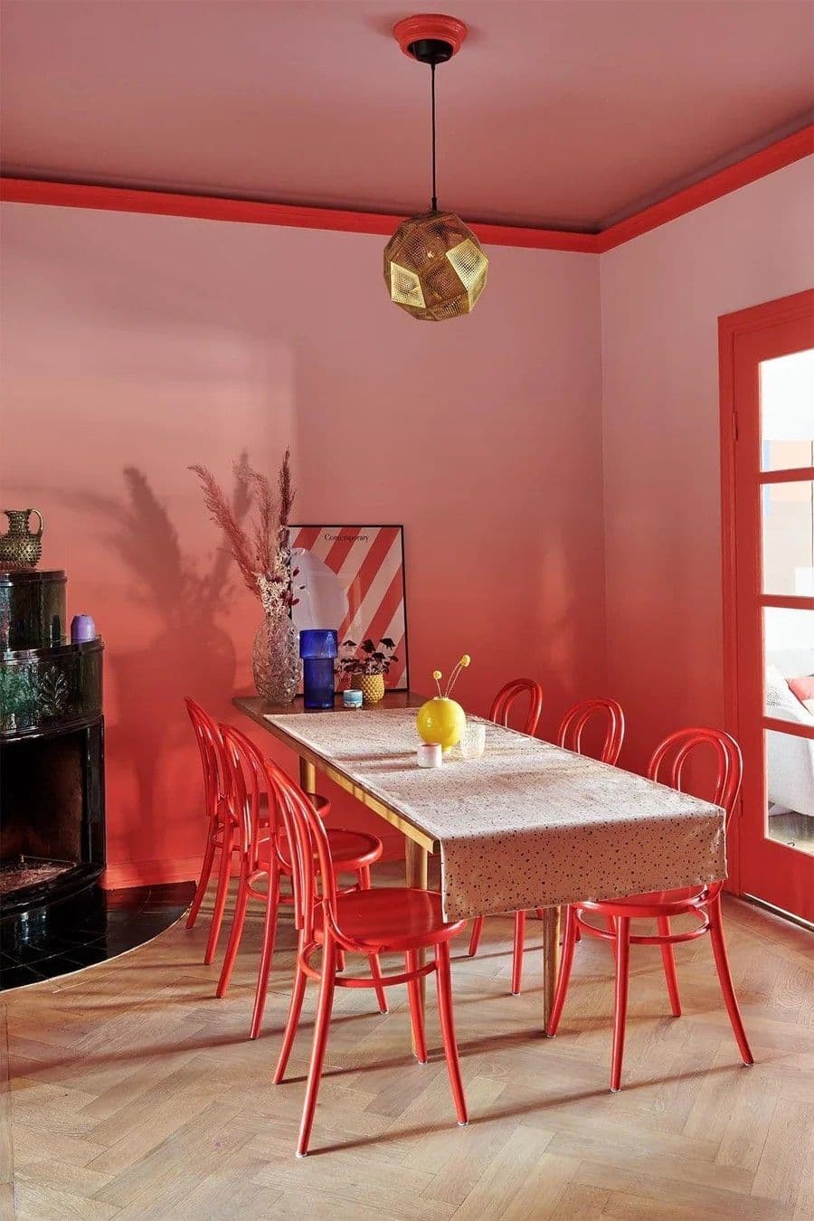 Comedor con muros pintados rojos, estilo ombré o degradado. Gran mesa de madera, rectangular, para 8 personas. 7 sillas de madera roja. Lámpara de techo, geométrica de metal dorado. Piso de parqué de madera clara. En la esquina hay una chimenea negra