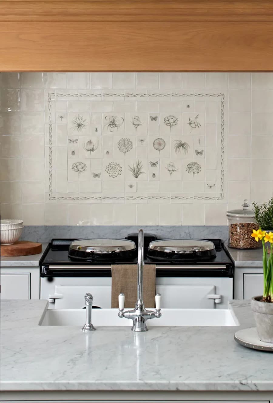 Cocina clásica con muro con azulejos color beige verdoso. Al centro hay un diseño con azulejos pintados mano con flores, mariposas y hojas. Muebles de cocina blancos, grifería cromada estilo vintage.
